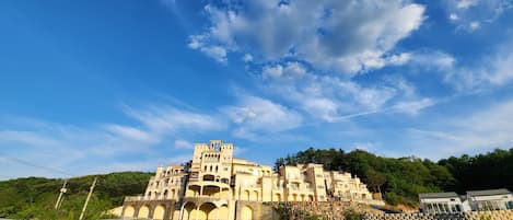 A large, multi-story building with arched windows and balconies, set against a clear blue sky with scattered clouds.