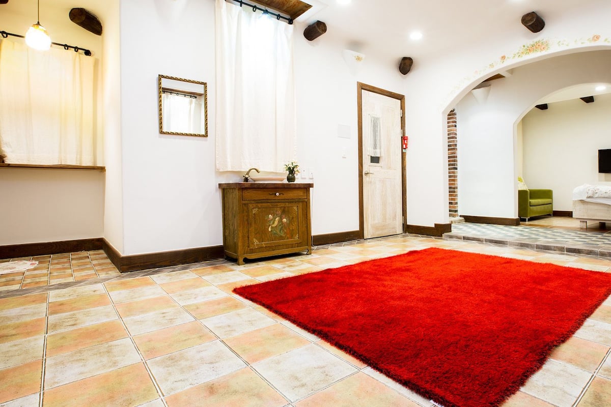 A hotel room with a red rug, a wooden cabinet, a mirror, a door, a TV, and a green sofa.