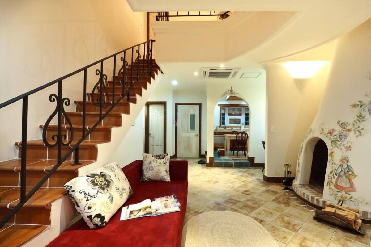 A spacious living area with a wooden staircase, a red sofa, and a kitchen in the background.