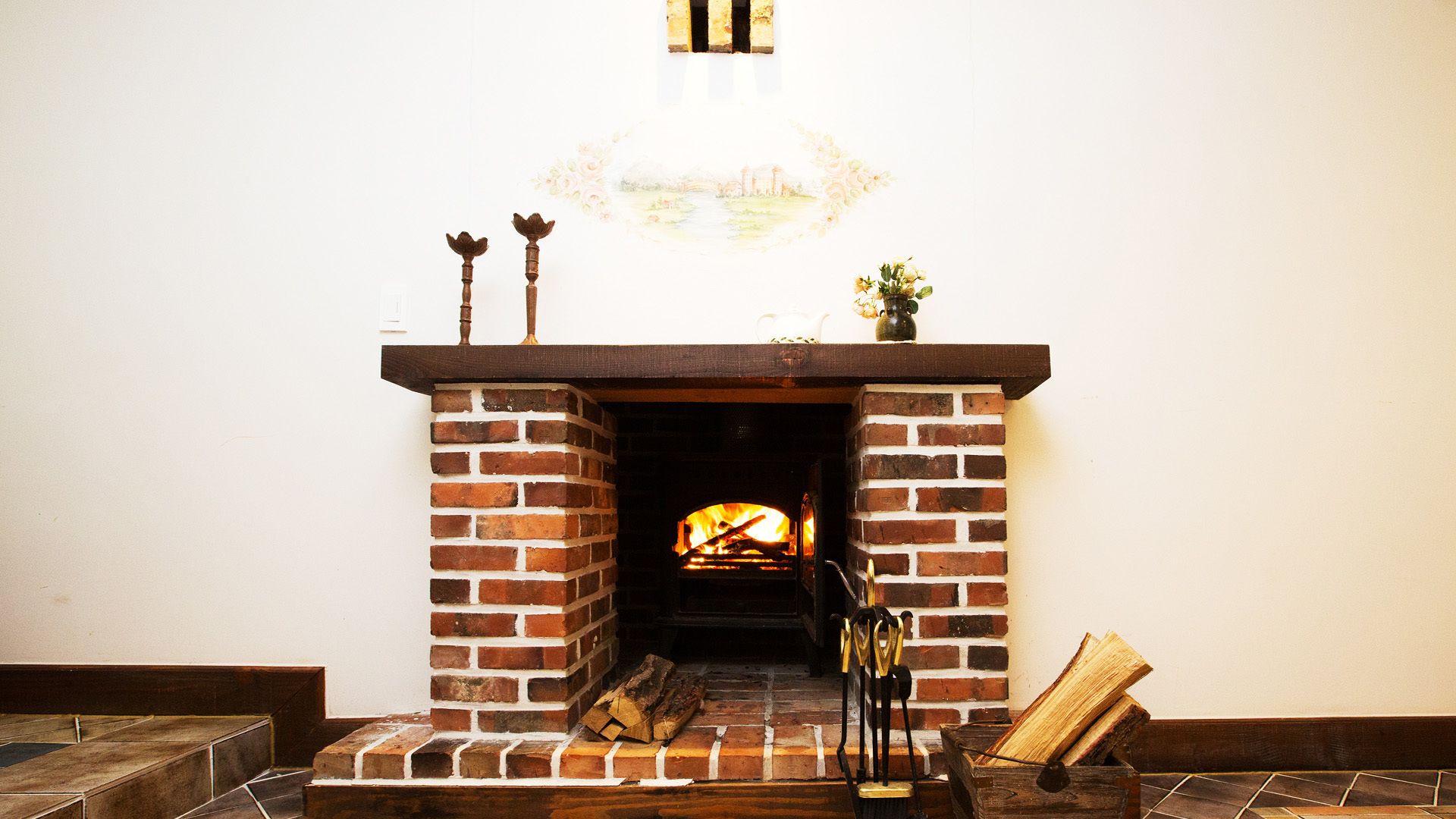 A brick fireplace with a fire, a vase with flowers, and a wooden box with logs.