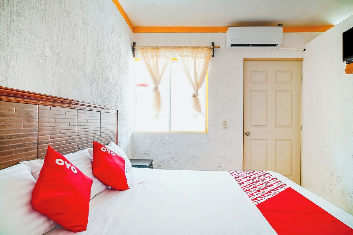 A hotel room with a wooden headboard, a bed with white and red bedding, a window with curtains, and an air conditioning unit.