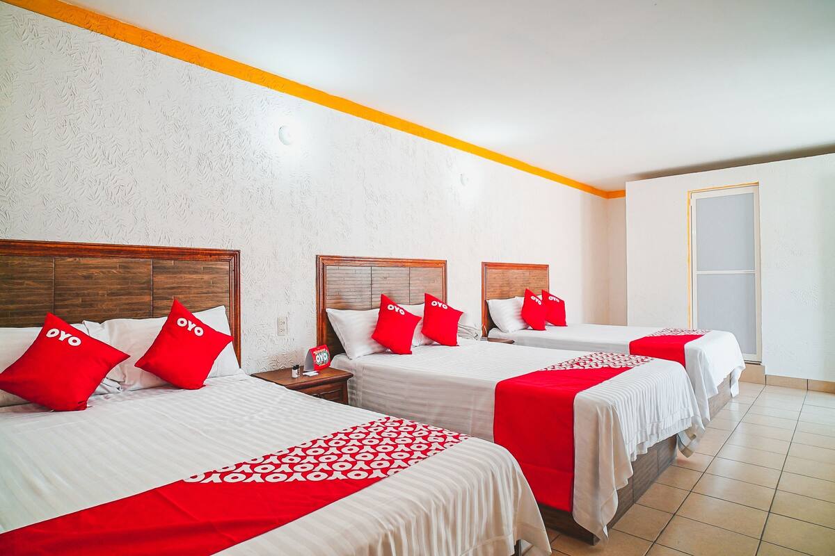 A hotel room with three beds, each with a wooden headboard, white bedding, and red accents.