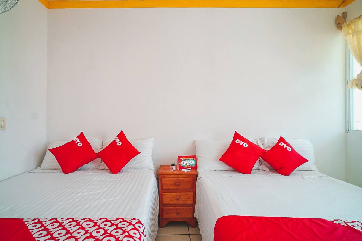 A hotel room with two beds, each with red and white bedding, a wooden nightstand with a box and a bottle, and a window with curtains.