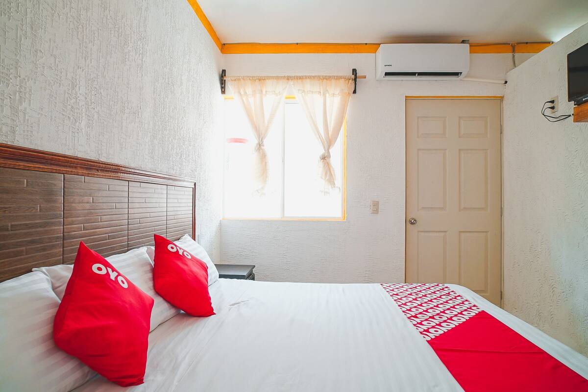 A hotel room with a bed, two red pillows, a white bedspread, a window with curtains, a door, and an air conditioning unit.
