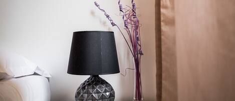 Eine moderne Lampe mit geometrischem Design auf einem Nachttisch neben einer Vase mit violetten Blumen.