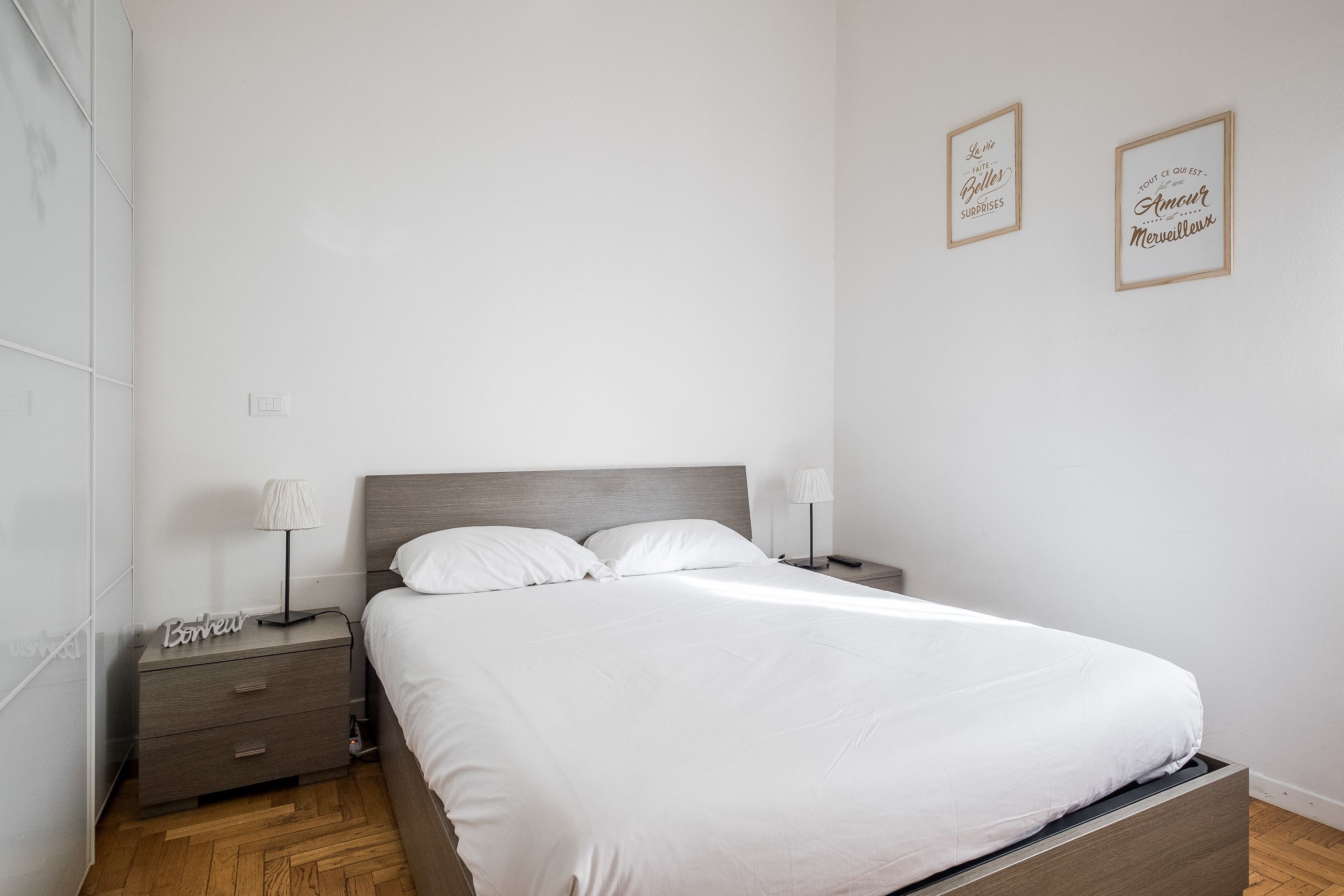 Una cama bien hecha en una habitación con cabecera de madera, dos mesitas de noche con lámparas y dos citas inspiradoras enmarcadas en la pared.