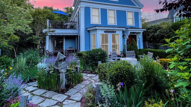A two-story house with a blue exterior, a front porch, and a garden with various plants and flowers. - Stylish Dog-Friendly Condo - West End on Comm St (Provincetown)