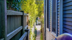 A narrow pathway flanked by wooden fences and vegetation, leading to a white picket fence and a house with blue siding. - Stylish Dog-Friendly Condo - West End on Comm St (Provincetown)
