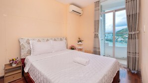 A bedroom with a bed, a nightstand with a flower vase, a window with curtains, and an air conditioning unit. - Cozzy Seaview Apartment (Ho Chi Minh City)