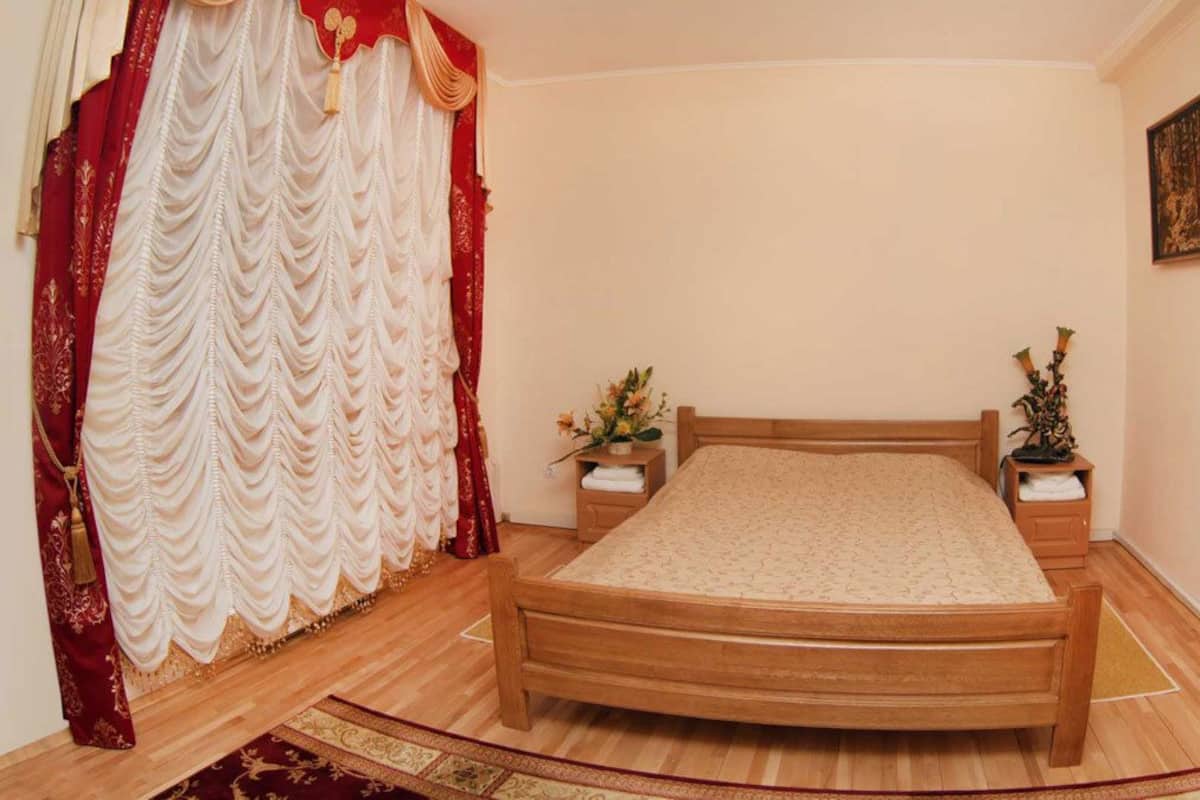 Un dormitorio con una cama de madera, una mesita de noche con lámpara, un cuadro en la pared y una cortina roja.