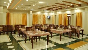 A dining area with multiple tables and chairs, a television mounted on the wall, and a ceiling with recessed lighting and wooden beams. - Hotel Shrinathinn (Nathdwara)