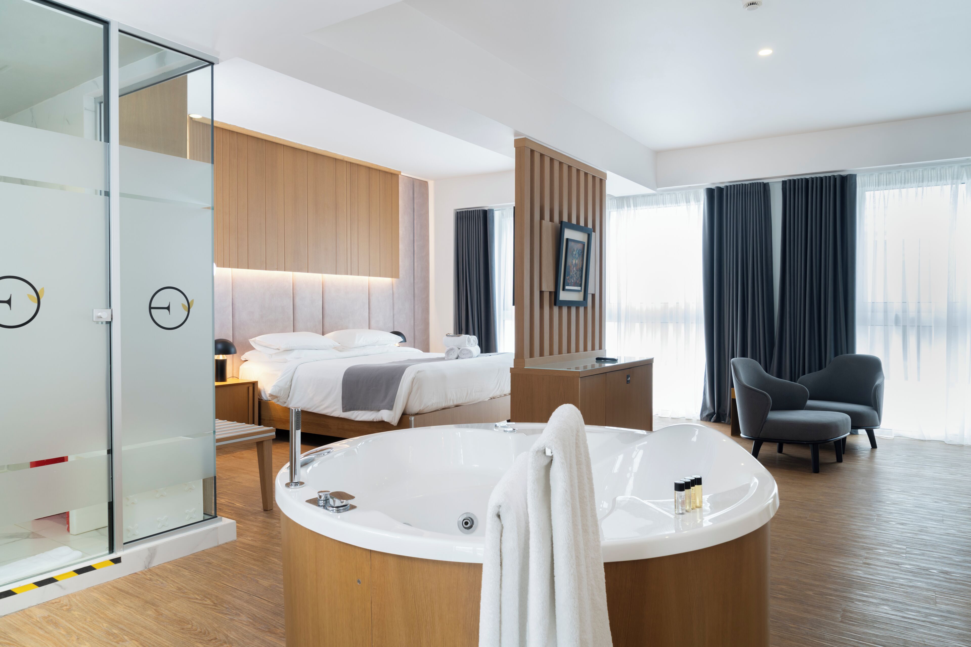 Una habitación de hotel moderna con una bañera grande, una cama con cabecero de madera, un box de ducha de vidrio y una zona de estar con una silla.
