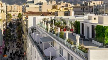 A modern hotel with a glass facade, outdoor seating, and a view of the city and mountains.