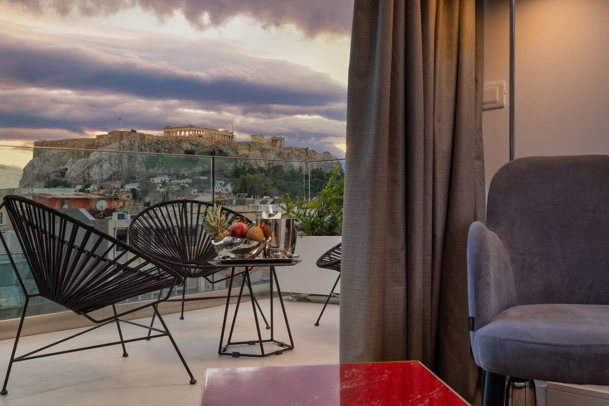 A balcony with a view of ancient ruins, two chairs, a table with fruits and a pitcher, and a red table.