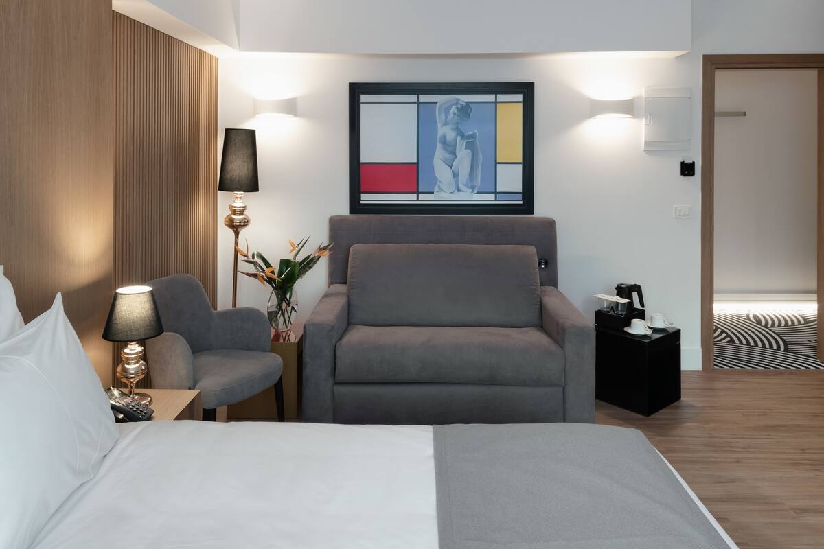 A modern hotel room with a large bed, a grey armchair, a small side table with a lamp, a framed artwork on the wall, and a black nightstand with a coffee maker.