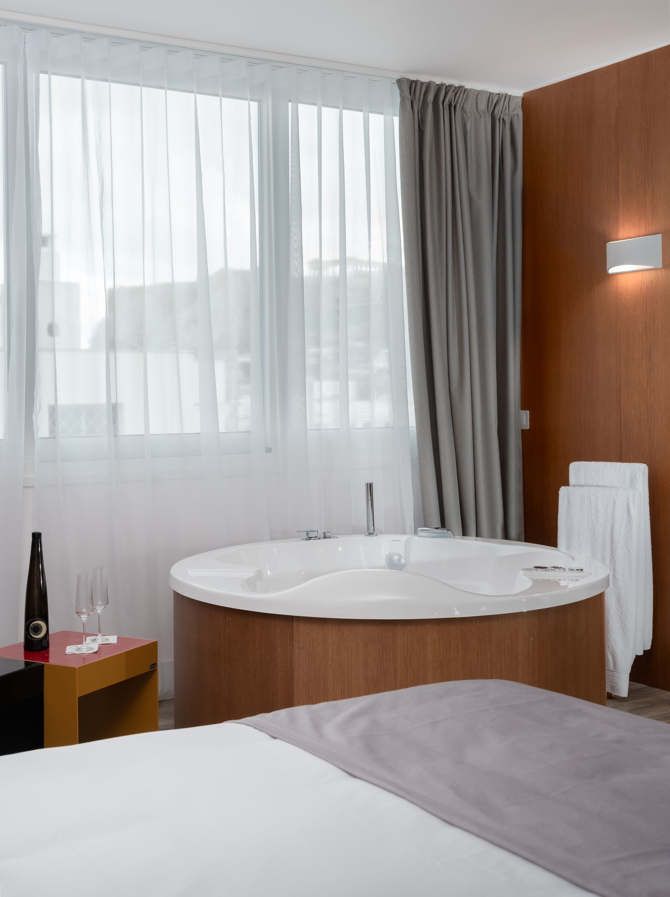 Une salle de bain d’hôtel moderne, dotée d’une grande baignoire, d’un meuble-lavabo en bois et d’une grande fenêtre agrémentée de rideaux légers.