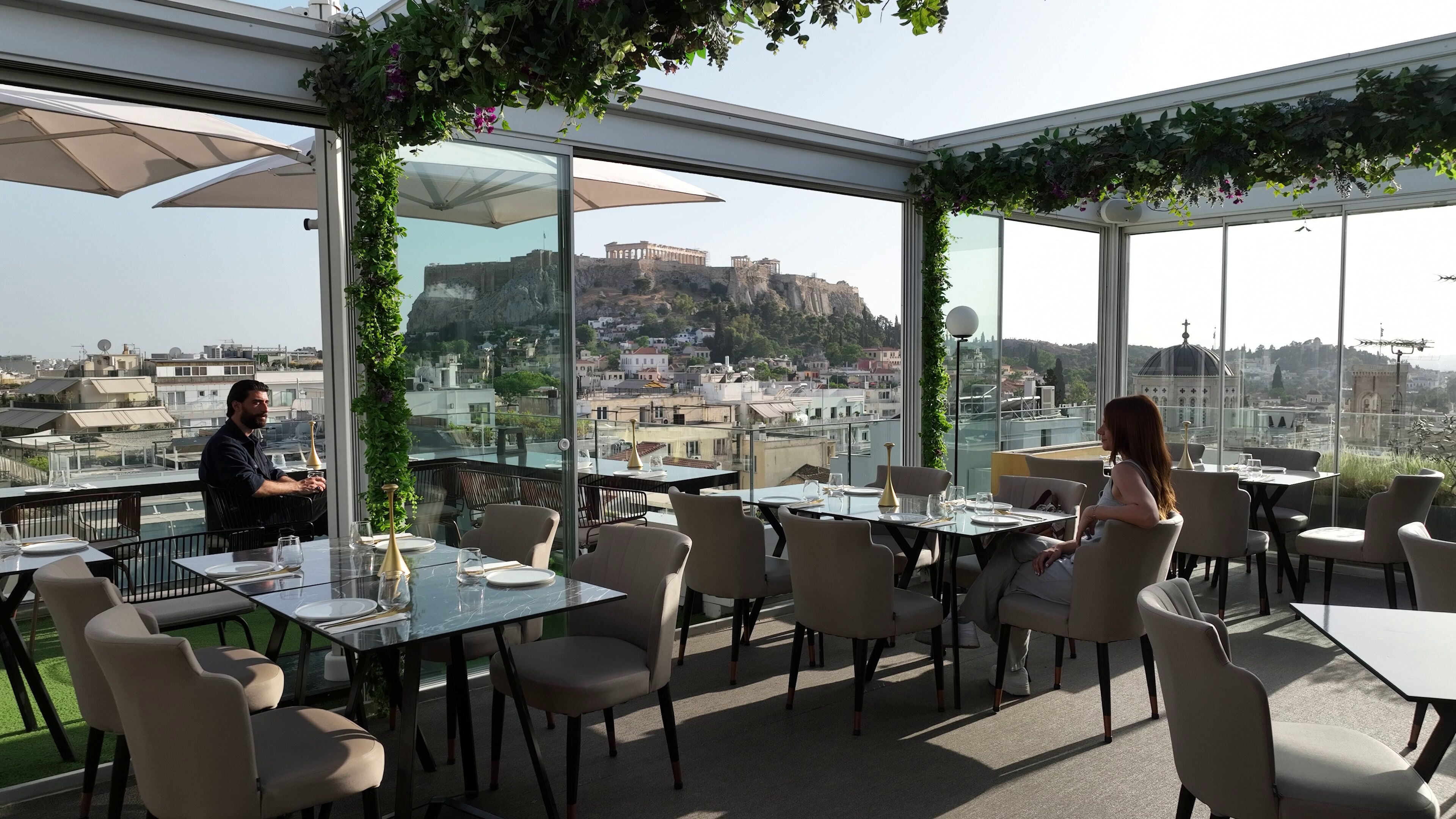 A rooftop dining area with tables and chairs, large windows offering a city view, and greenery hanging from the ceiling.