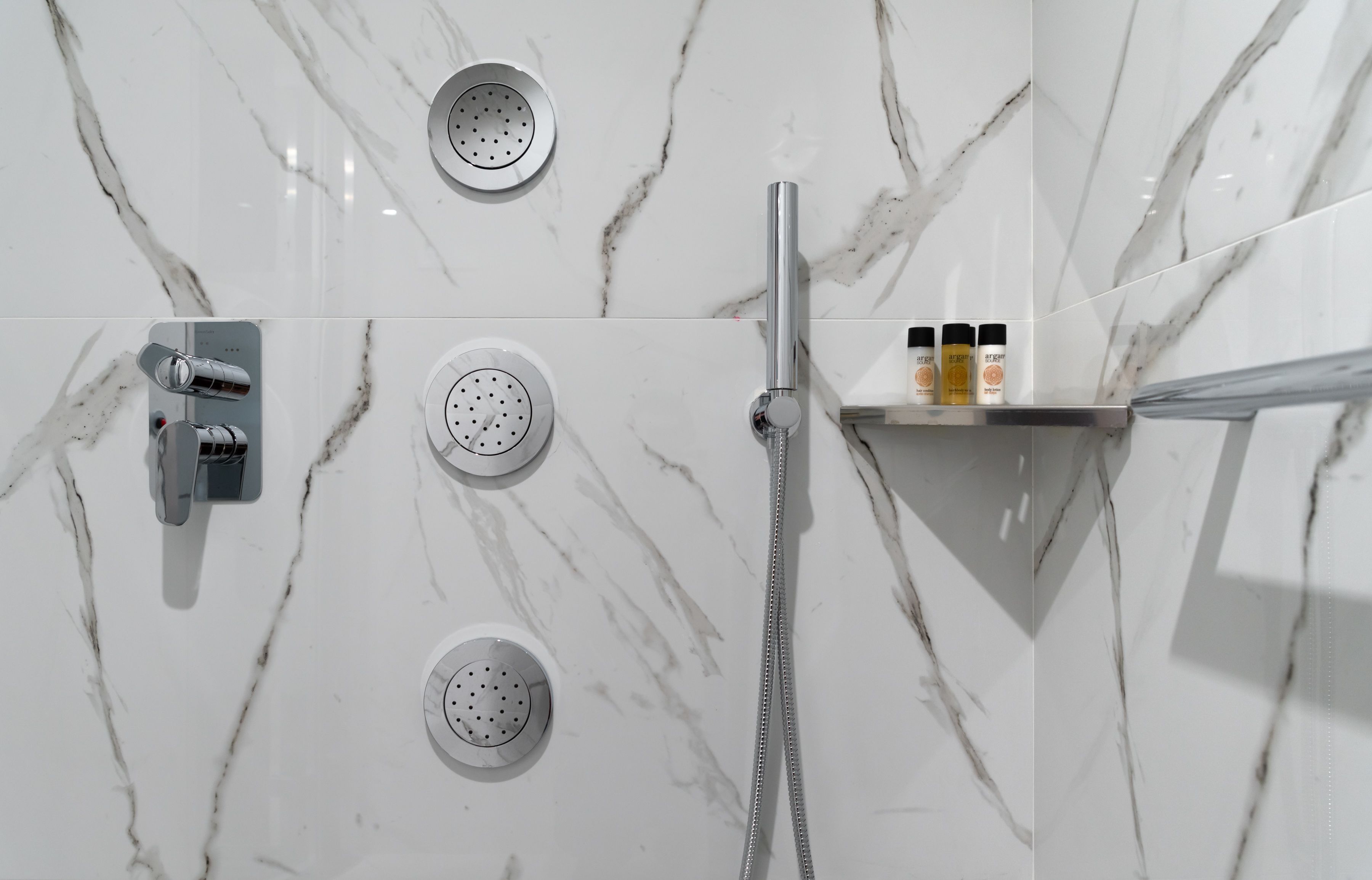 A bathroom with a marble wall, a handheld showerhead, and a shelf with toiletries.
