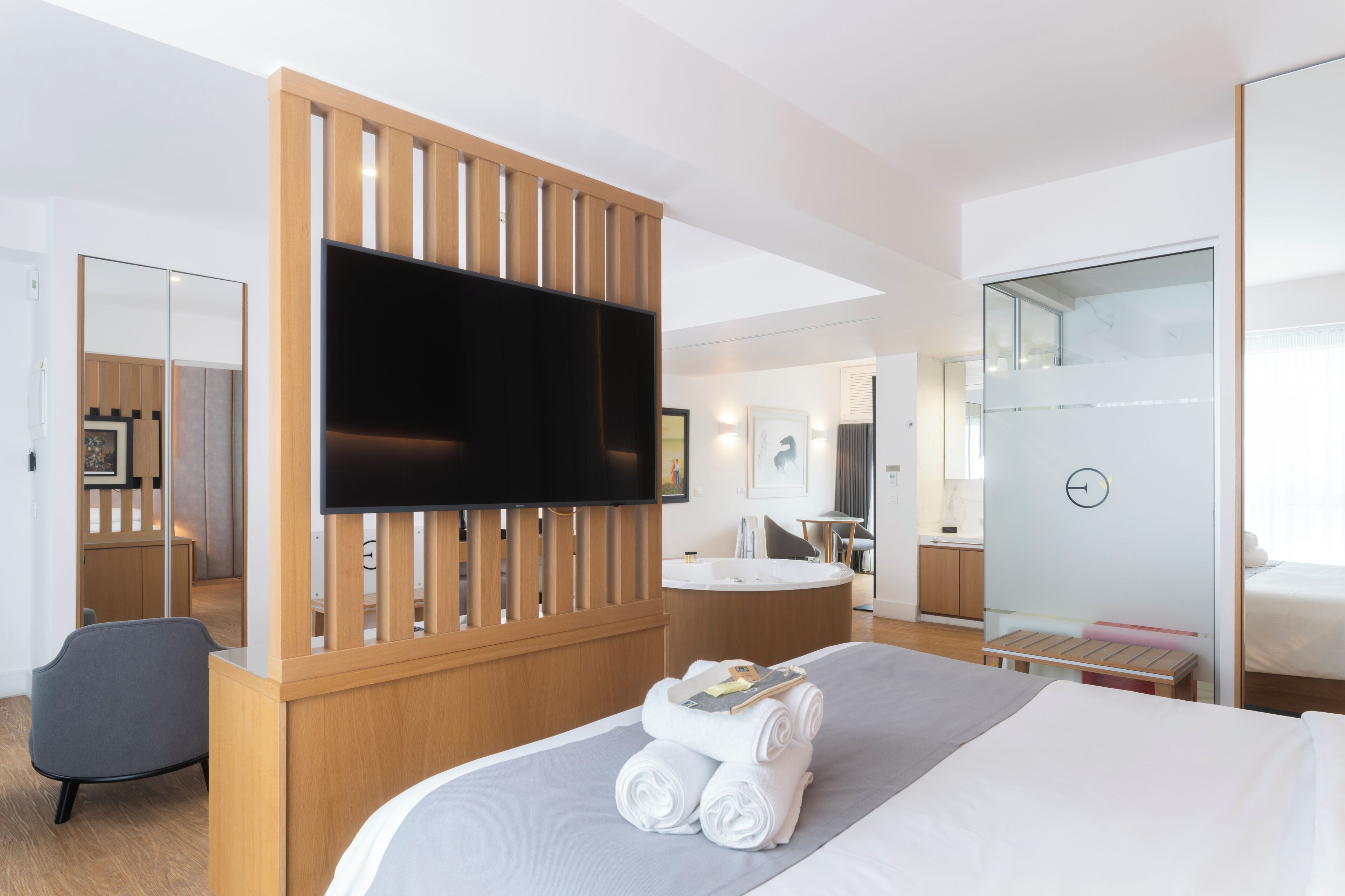 Una habitación de hotel moderna con una cama grande, un televisor de pantalla plana, un separador de madera, una zona de estar y un baño con puerta de vidrio.