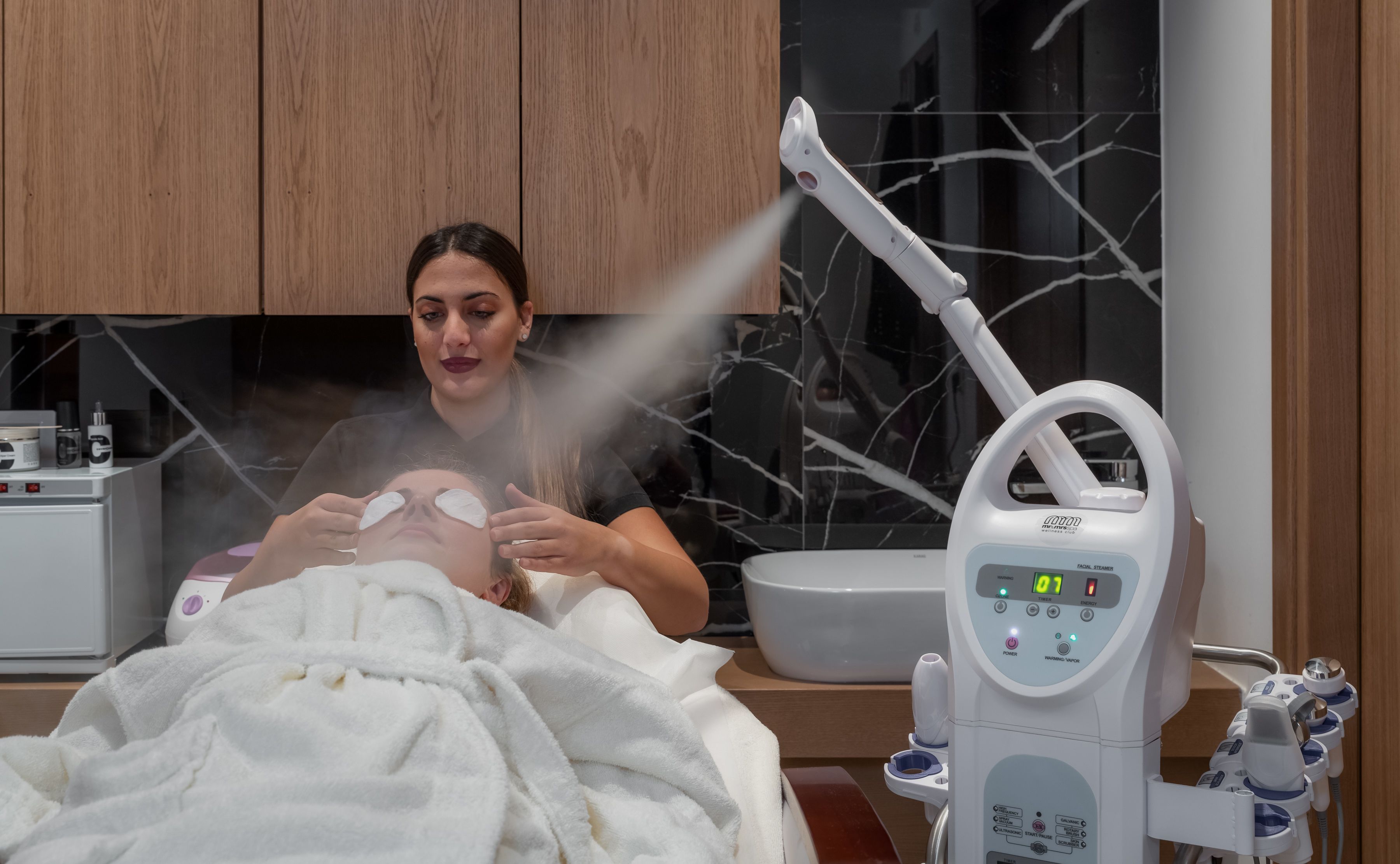 A facial steam treatment in a spa setting with a steam machine and a person applying a facial mask.