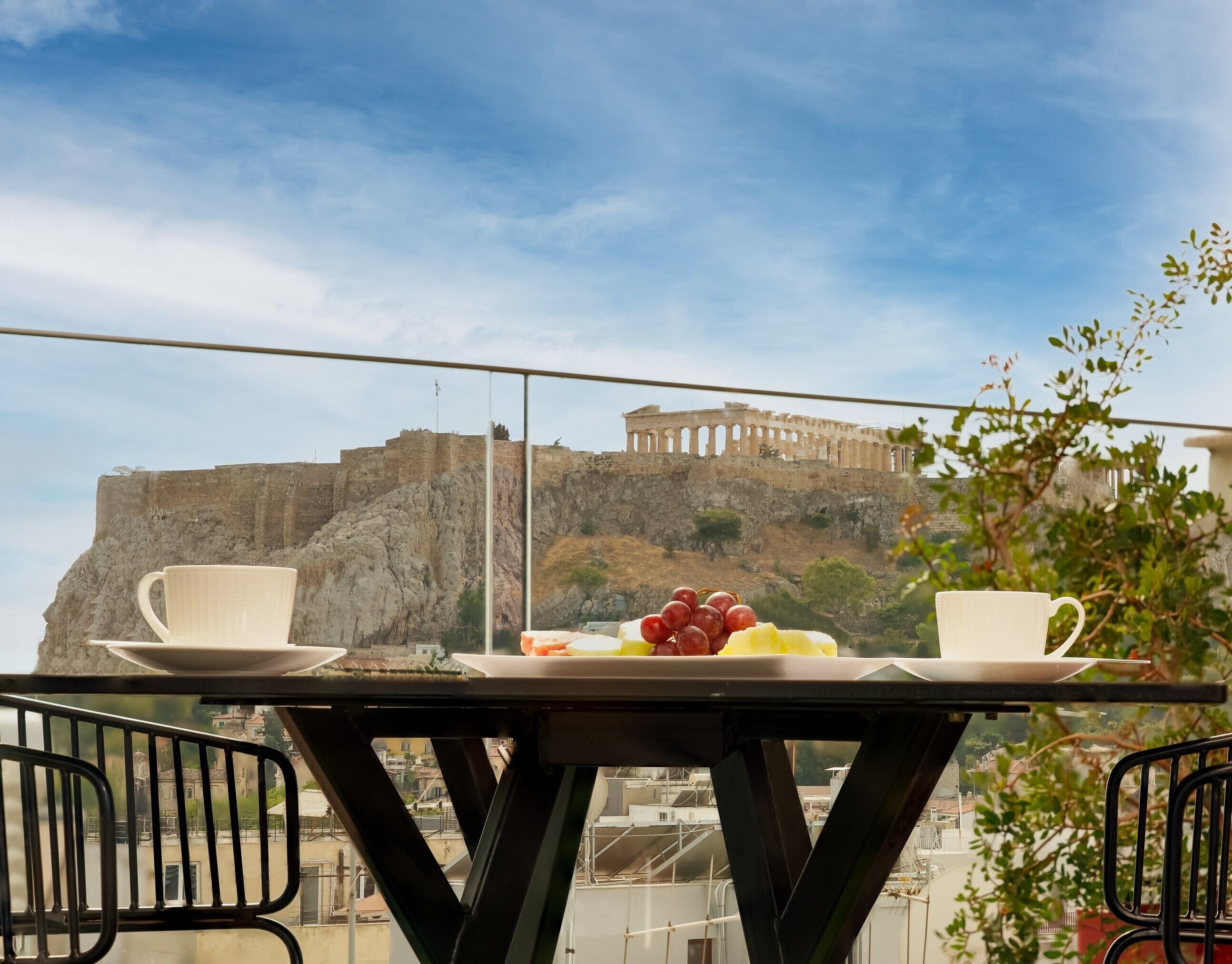 A balcony table with a white plate of fruit and two white cups, overlooking a historic stone fortress with ancient architecture.