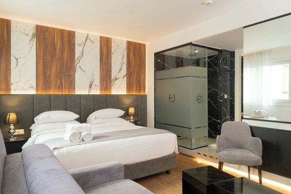 Una habitación de hotel moderna con una cama grande, un sofá, un vestidor amplio con paredes de vidrio y un baño con un espejo grande.