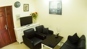 Een woonkamer met twee zwarte banken, een salontafel, een televisie op een witte kast, een klok en een ingelijst kunstwerk aan de muur.