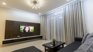 A modern living room with a flat-screen TV mounted on a wooden panel wall, a dark sofa, a coffee table, and a large window with curtains. - Cozzy and Beauty Apartment (Ho Chi Minh City)