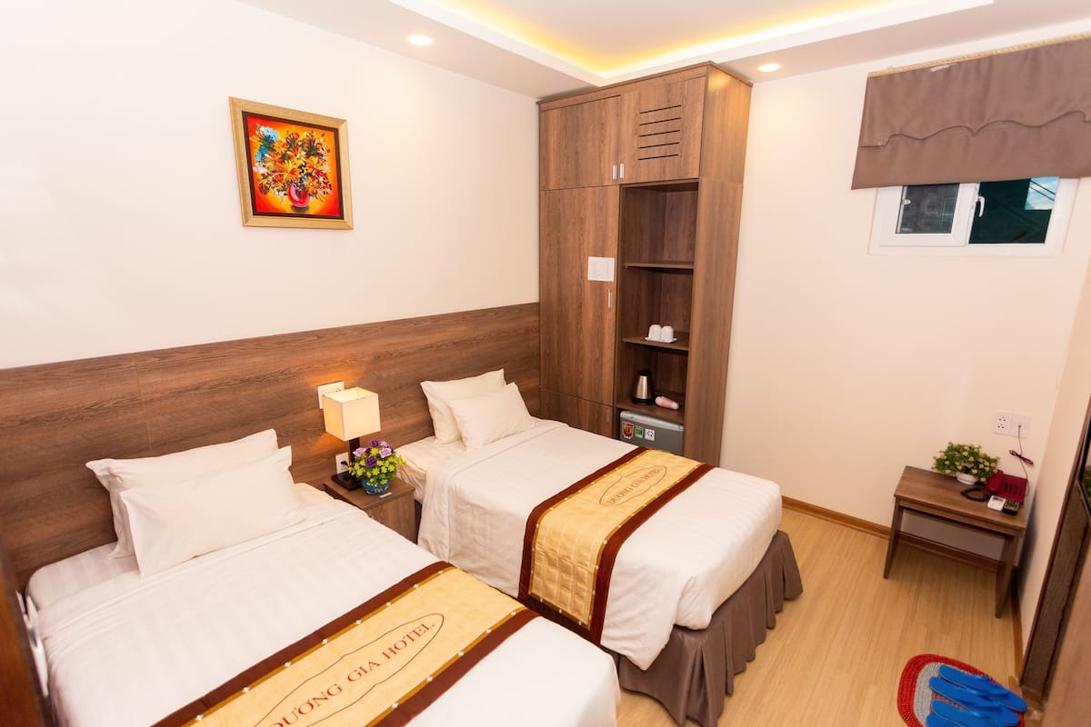 Une chambre d’hôtel avec deux lits, une armoire, une petite table avec une lampe et une fenêtre avec des rideaux.