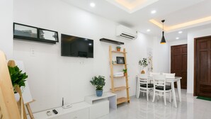 Un salon moderne avec un téléviseur à écran plat, des meubles blancs, une étagère en bois en forme d’échelle, une table à manger avec des chaises et une plante en pot.