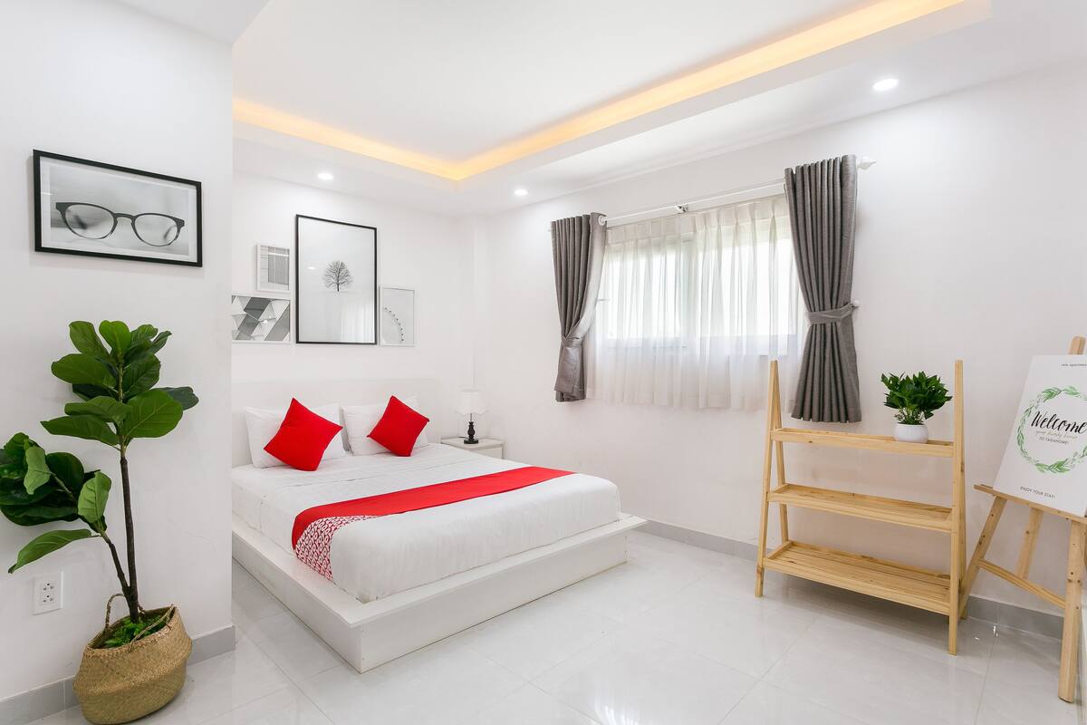 A modern bedroom with a white bed, red pillows, a small potted plant, and a welcome sign.