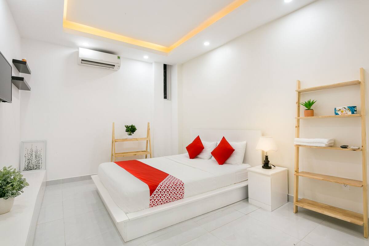 A modern hotel room with a bed, bedside table, lamp, air conditioning unit, and a wooden shelf with a plant.