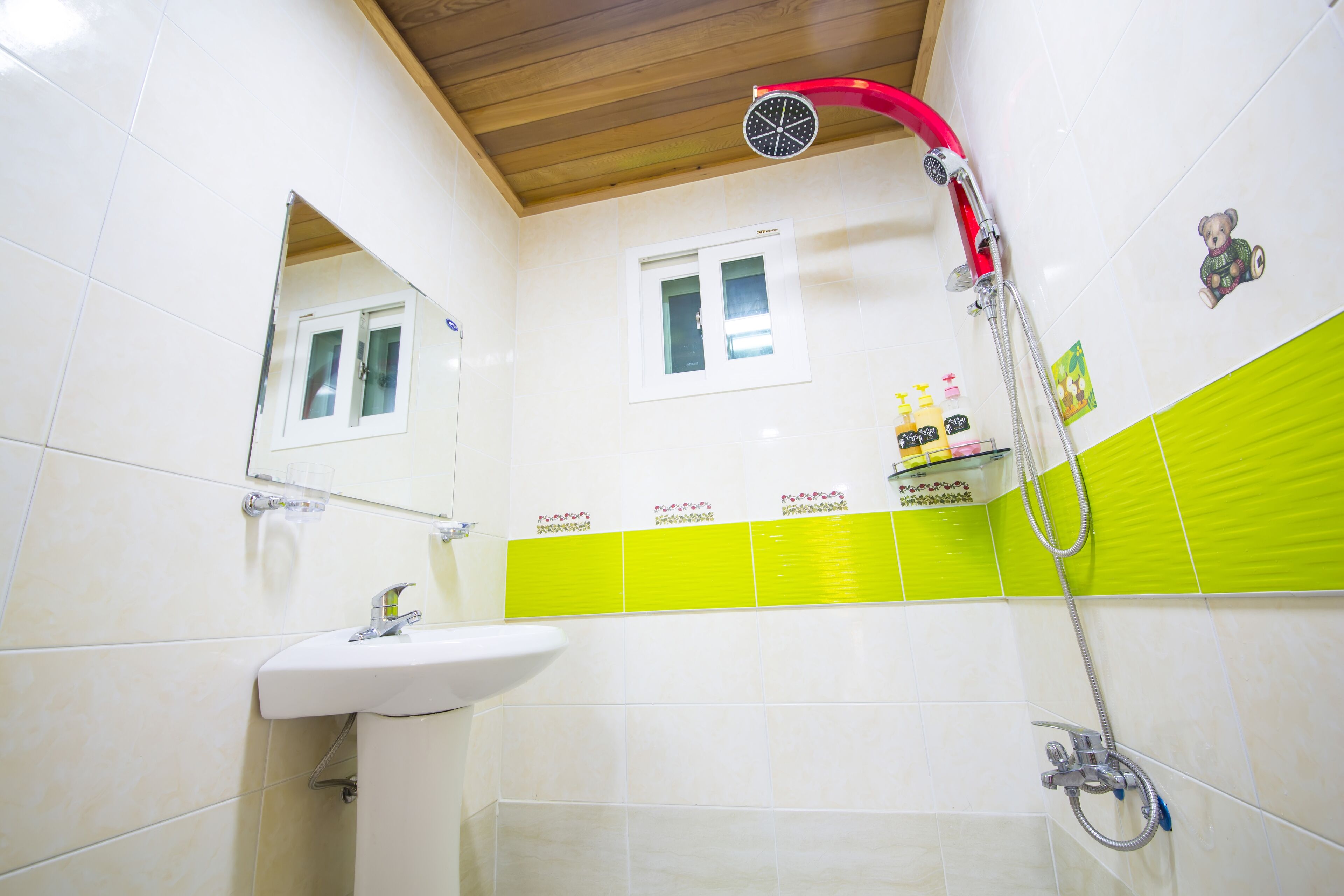 Un baño con lavamanos blanco, un espejo, una ducha con manguera roja y azulejos verdes.