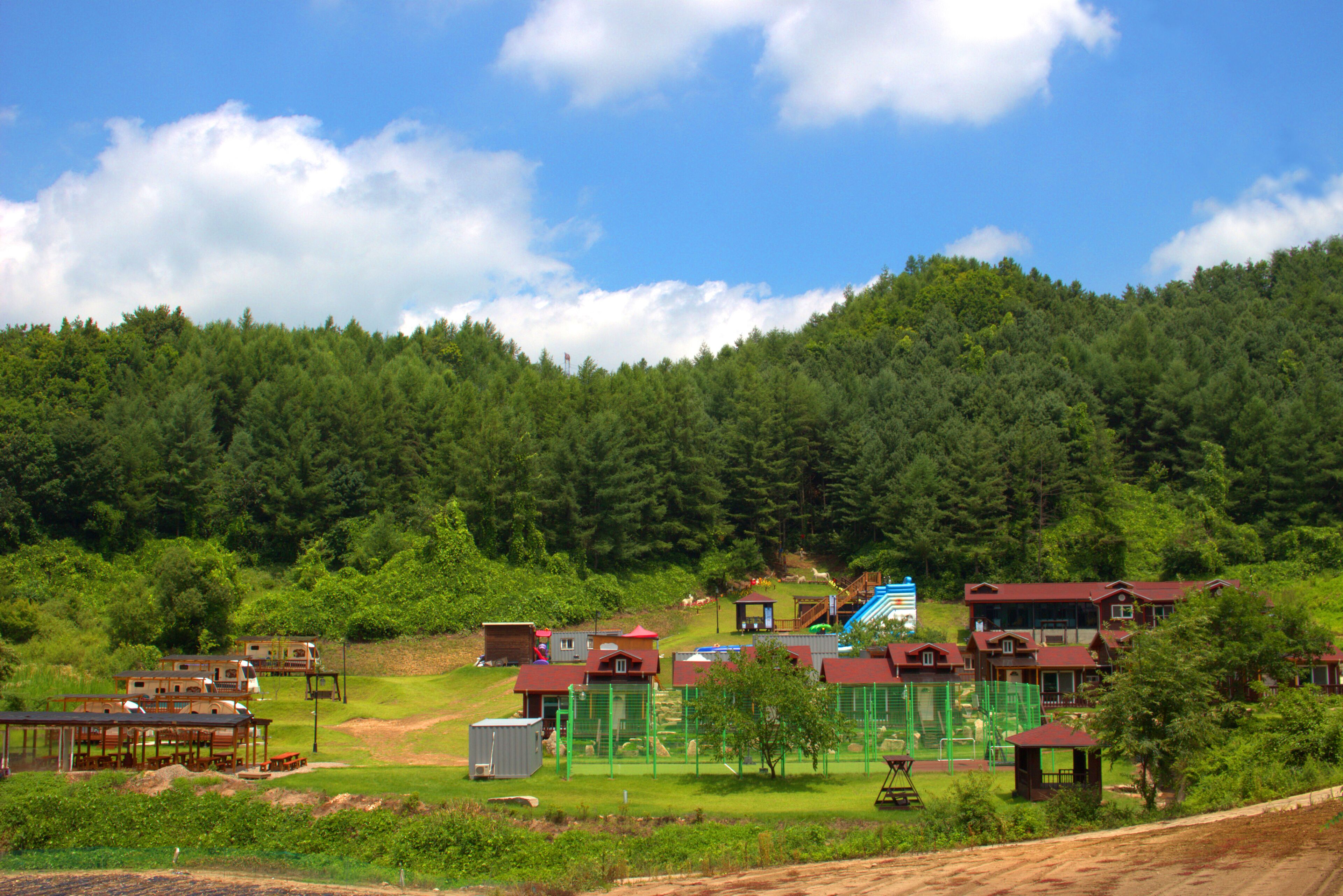A rural setting with multiple buildings, including cabins and a larger house with a red roof, surrounded by lush greenery and a forested hill in the background.