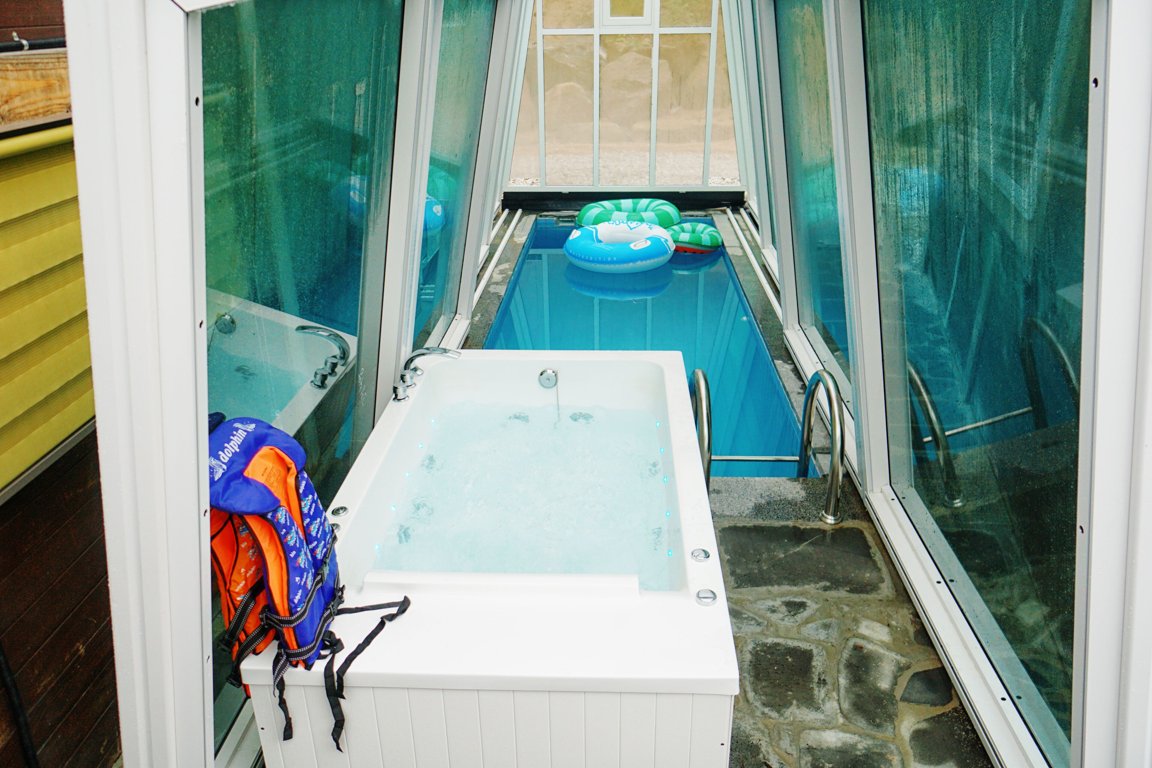 A small indoor pool with a hot tub, surrounded by glass walls and a window.