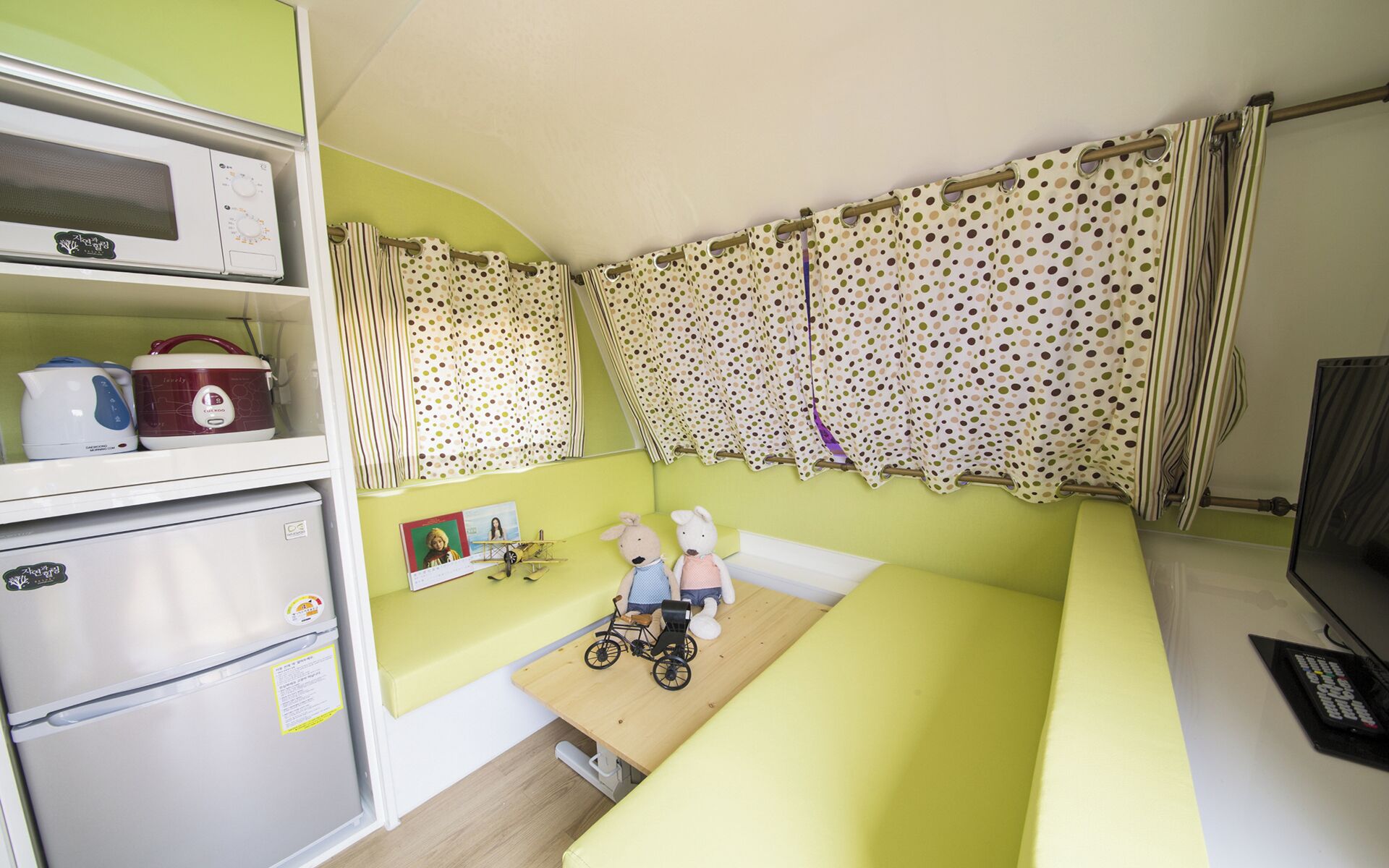 A compact room with a bed, a small table, a microwave, a refrigerator, and a window with polka dot curtains.