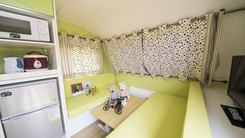 A compact room with a bed, a small table, a microwave, a refrigerator, and a window with polka dot curtains.