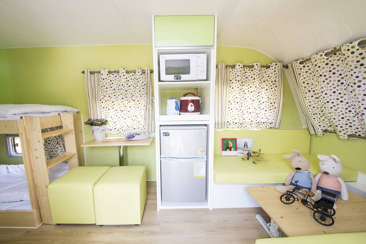 A bunk bed, a small kitchenette with a microwave, refrigerator, and kettle, a table with toys, and a window with curtains.