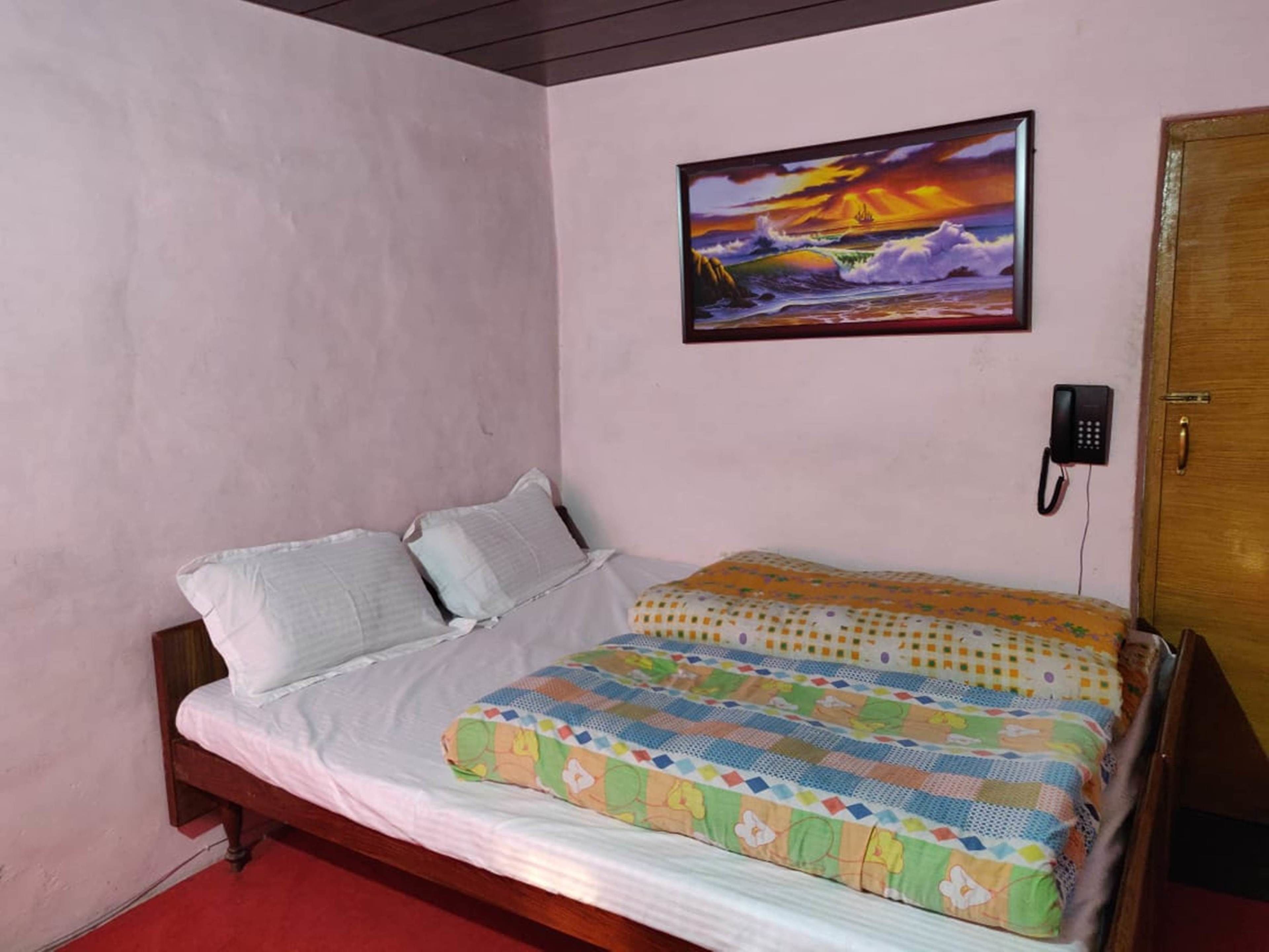 A single bed with a colorful quilt, a wooden headboard, a wall-mounted phone, and a painting of a sunset over the ocean.