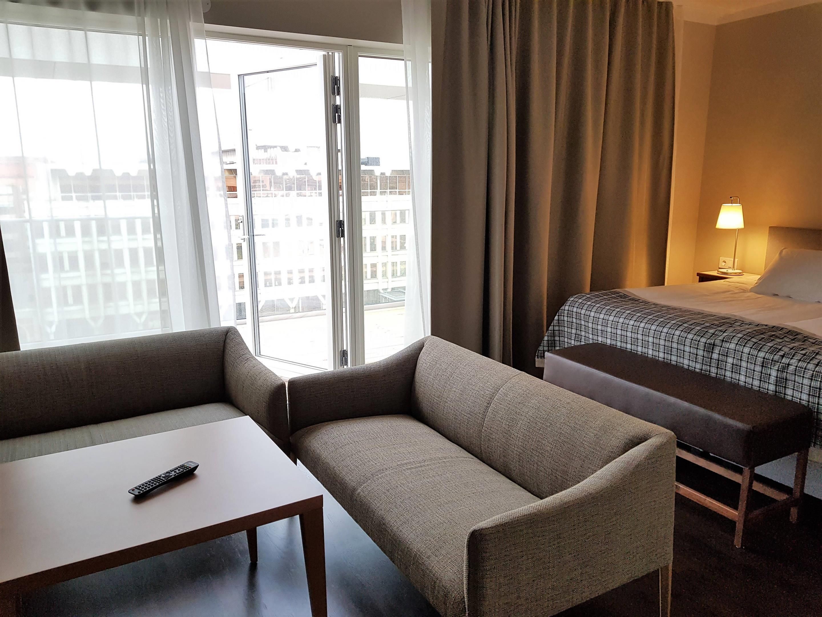 Ett hotellrum med en soffa, ett litet bord, en säng med ett rutigt överkast, ett sängbord med en lampa och utsikt över byggnader genom ett stort fönster.