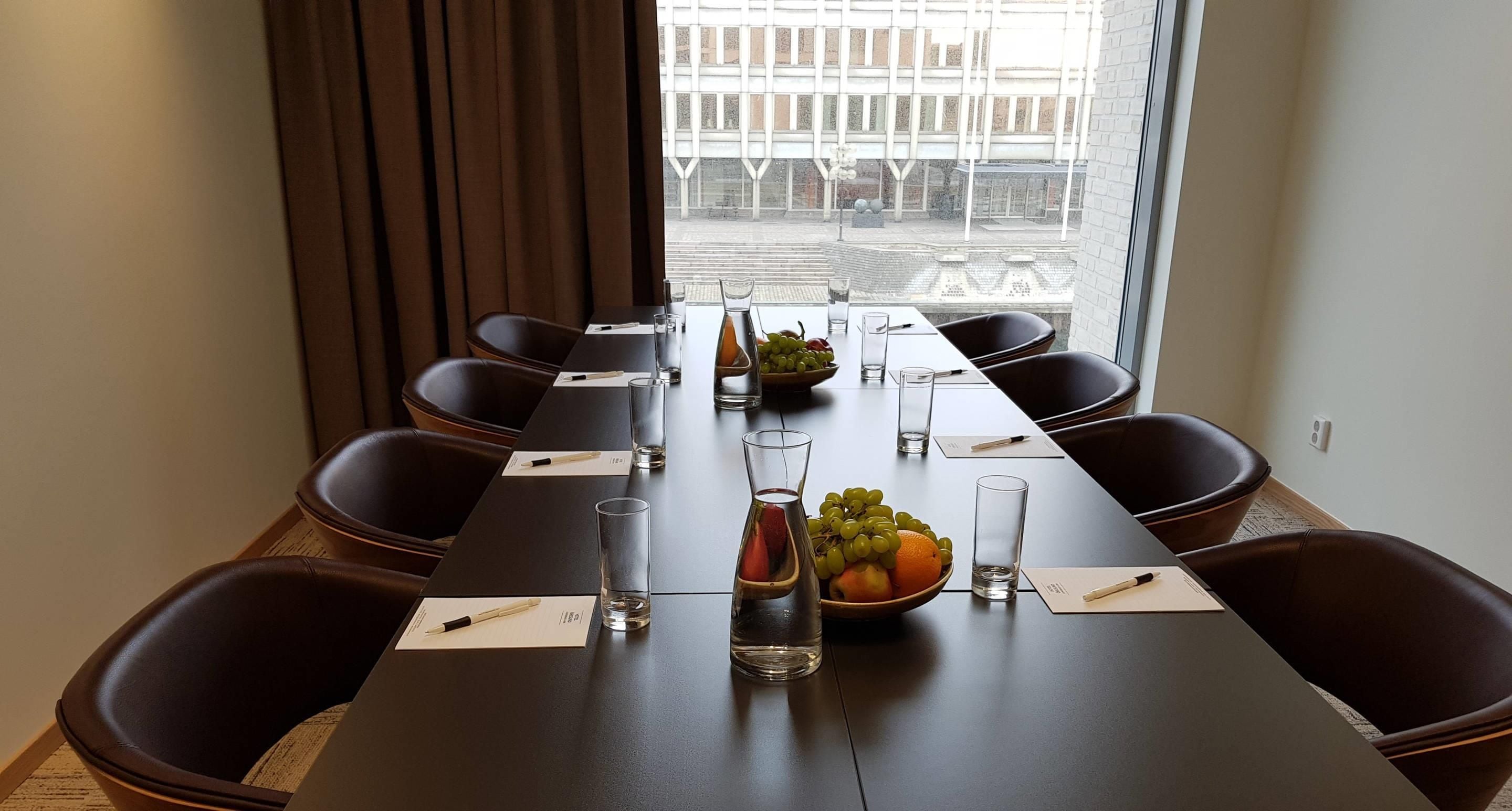 Ett konferensrum med ett långt bord, stolar, vattenglas, en fruktskål och ett fönster med utsikt över en byggnad.