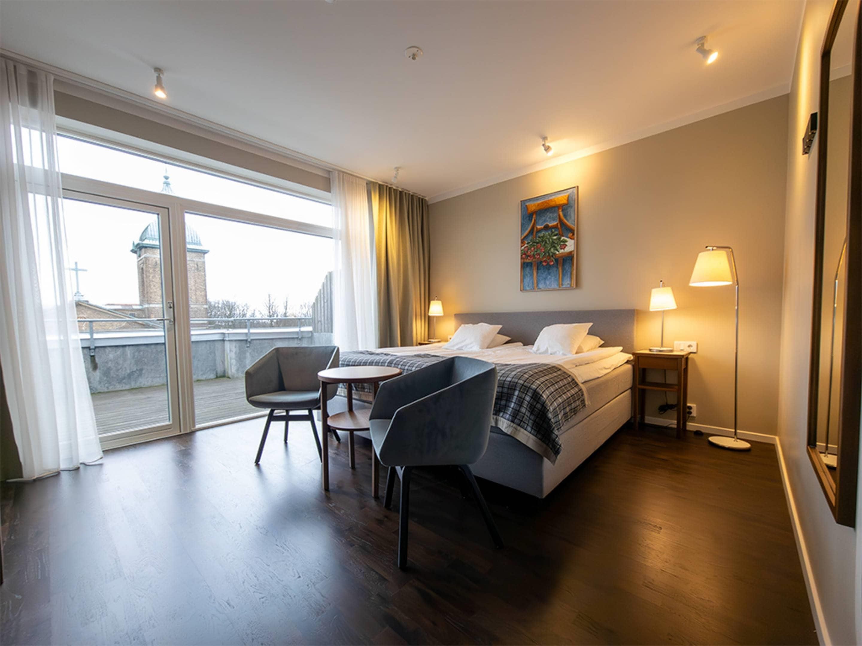 A hotel room with a bed, two bedside tables, a chair, a desk, and a large window with curtains.