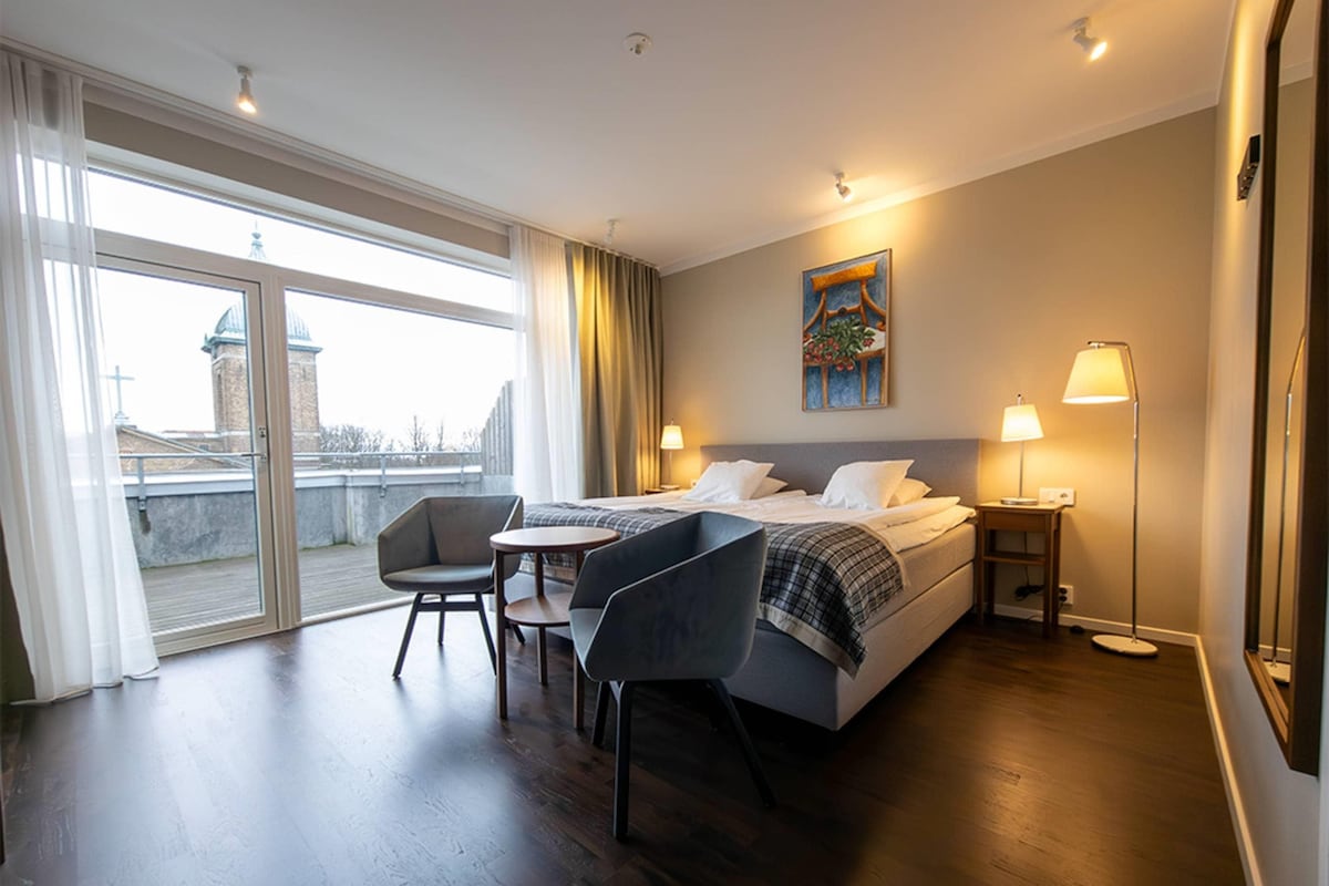 A hotel room with a bed, two bedside tables, a chair, a desk, and a large window with curtains.