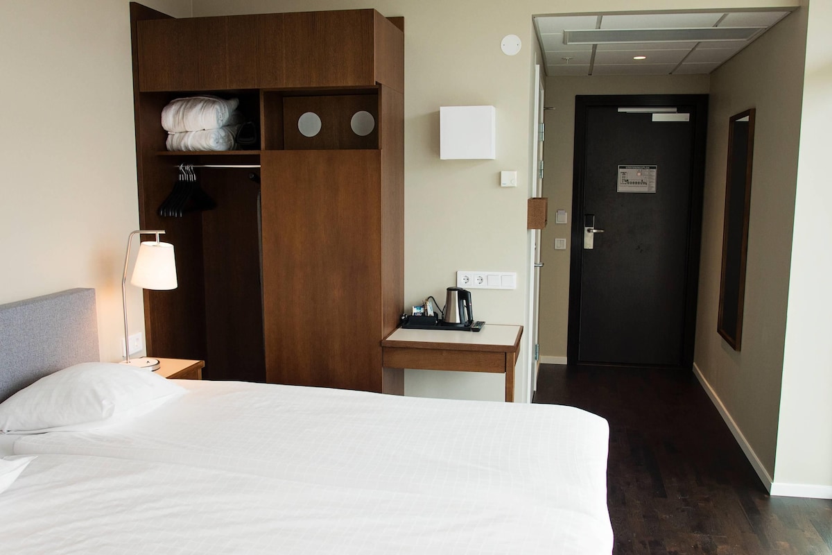 A hotel room with a bed, a desk, a wardrobe, a lamp, and a door leading to another room.