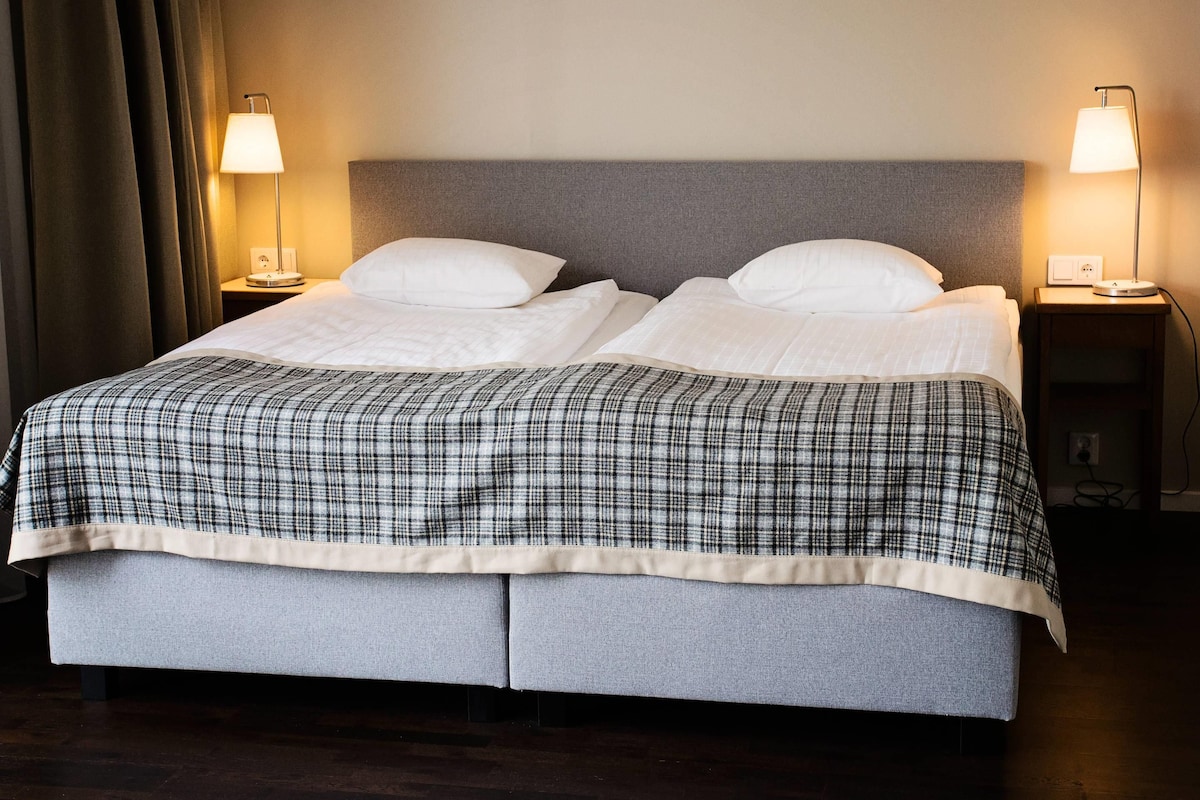 A double bed with a grey headboard, two bedside lamps, and a plaid bedspread.