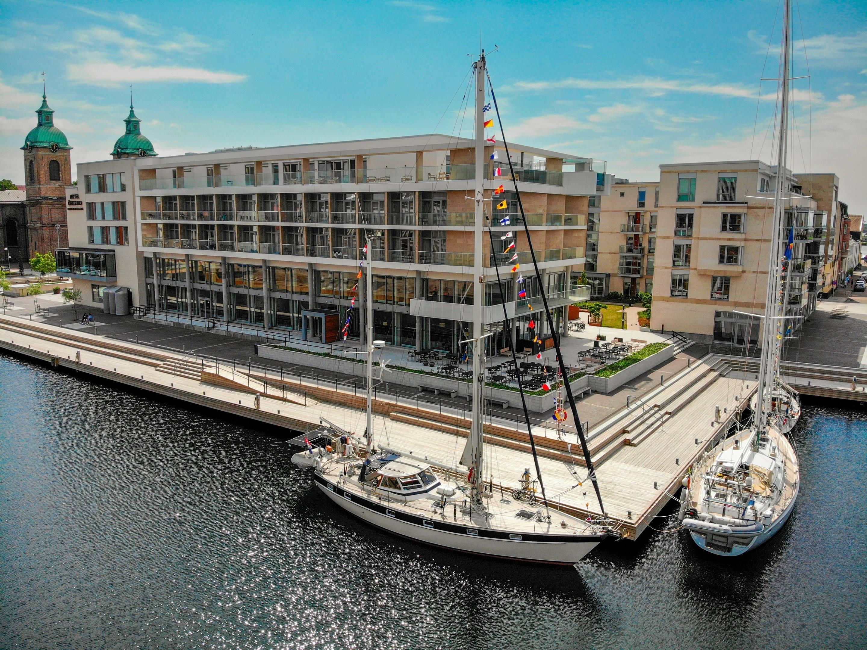 A modern building with multiple floors, a dock with a sailboat, and a historic tower in the background.