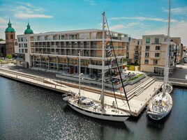 En modern byggnad med flera våningar, en brygga med en segelbåt och ett historiskt torn i bakgrunden.