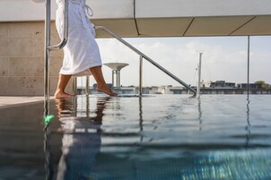 En person i en vit badrock som kliver ner i en pool med ett metallräcke, och i bakgrunden syns himlen och byggnaderna tydligt.