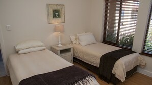 A bedroom with two beds, a nightstand, a lamp, and a window with blinds. - Villa 5 (Knysna)