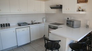 A modern kitchen with white cabinets, a dishwasher, a stove, a microwave, and a toaster. There are two black chairs at the island. - Villa 5 (Knysna)